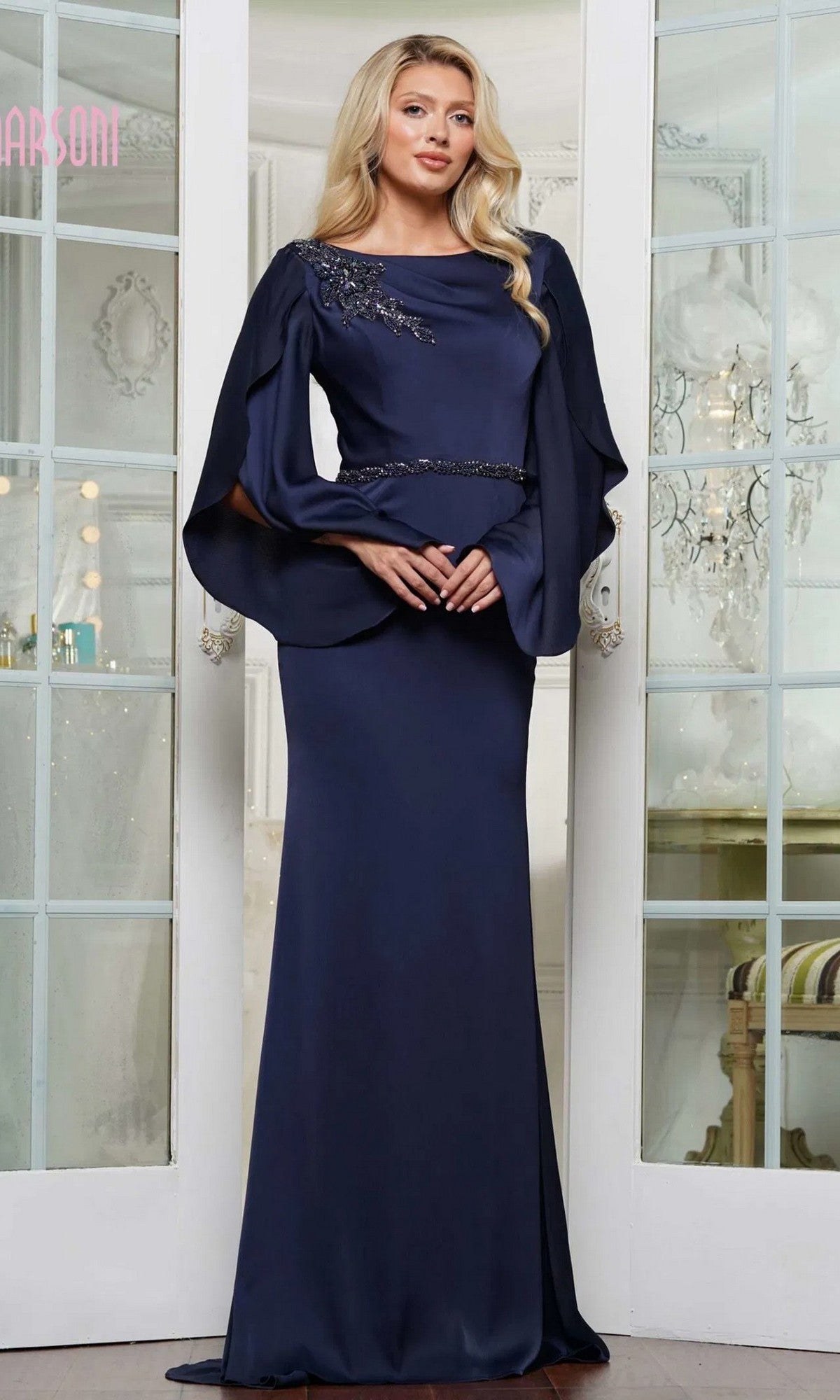Indoors, a woman wears the Marsoni MV1348 long formal dress in navy satin, showcasing beaded appliques on the shoulder and waist, and draped flowing sleeves.